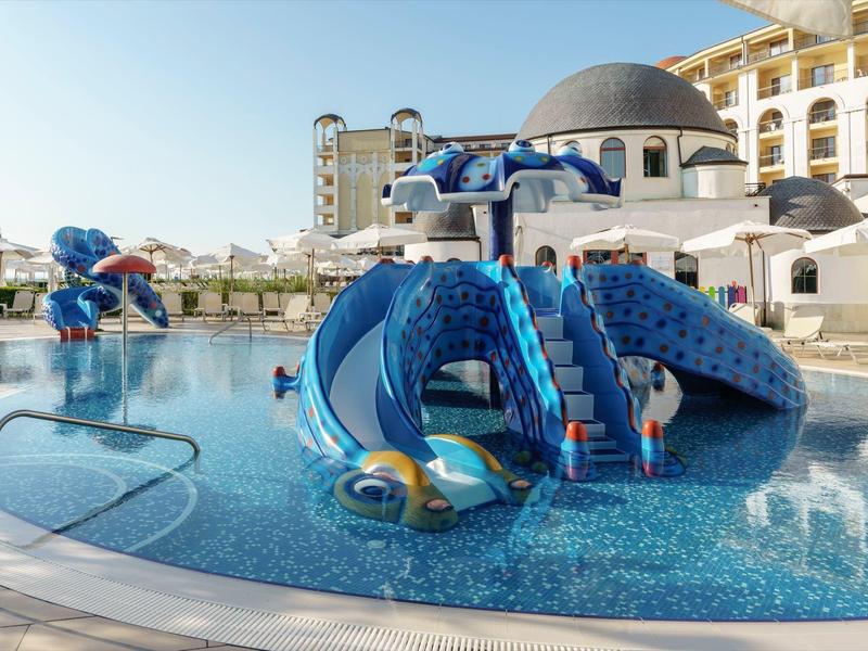 Hotelzwembad met blauwe glijbaan en speeltuin, parasols en gebouwen op de achtergrond.