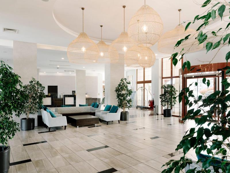 Lichte hotellobby met zithoeken, grote planten en moderne hanglampen.