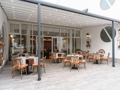 Überdachte Terrasse eines Restaurants mit Holztischen und Stühlen vor großen Fenstern.