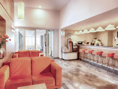 Lichte hotellounge met oranje bank, barkrukken en grote ramen die daglicht binnenlaten.