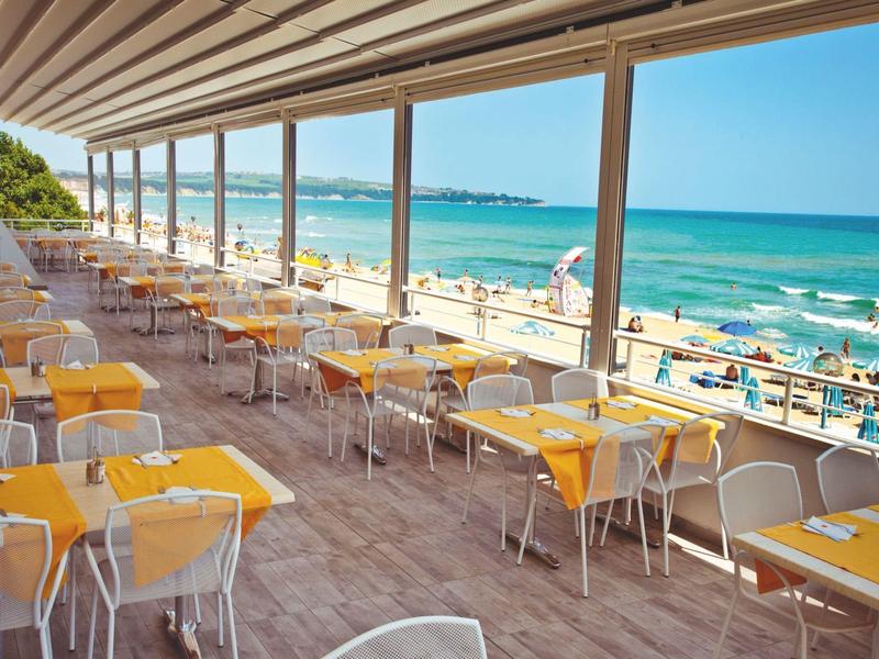 Restaurantterras met gele tafels met uitzicht op een druk strand en blauwe zee.