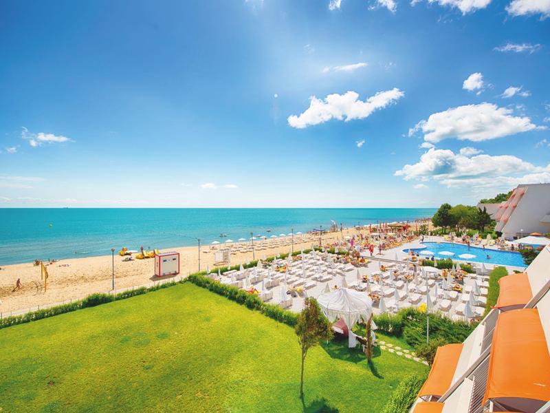 Uitzicht op een strand met parasols, ligstoelen en groen gras onder een blauwe lucht.