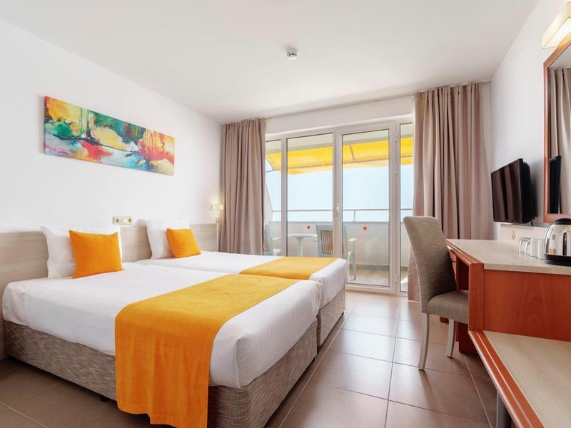 Modern hotelkamer met tweepersoonsbed, oranje bedsprei en balkonuitzicht.