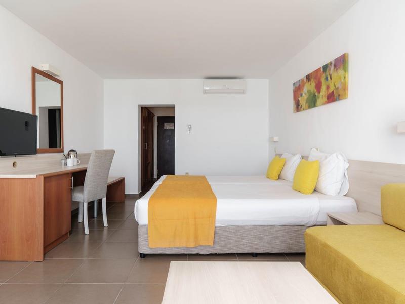 Lichte hotelkamer met tweepersoonsbed, gele accenten en een bureau.