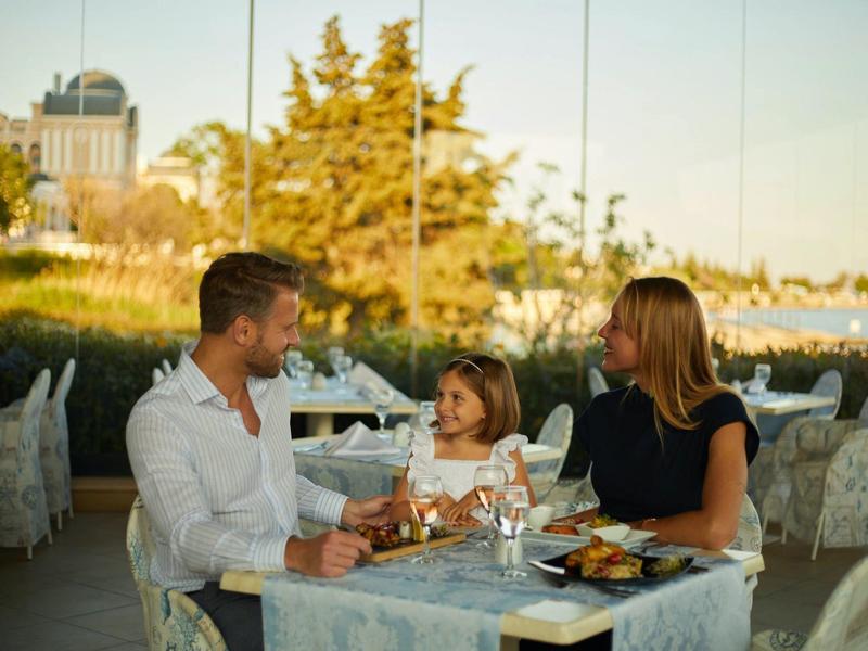 Family with child enjoying dinner and wine on terrace overlooking trees and buildings.