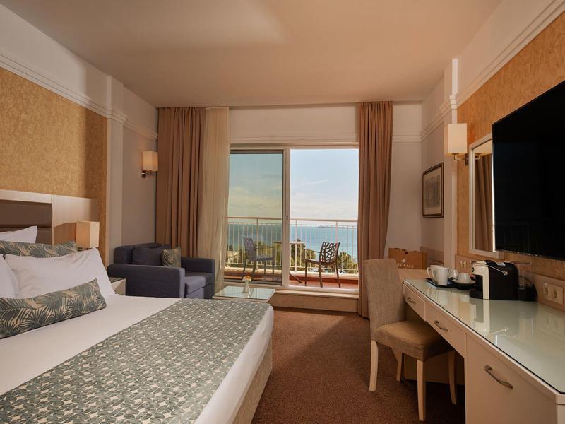 Modern hotel room with double bed, seating area, and sea view through large windows.
