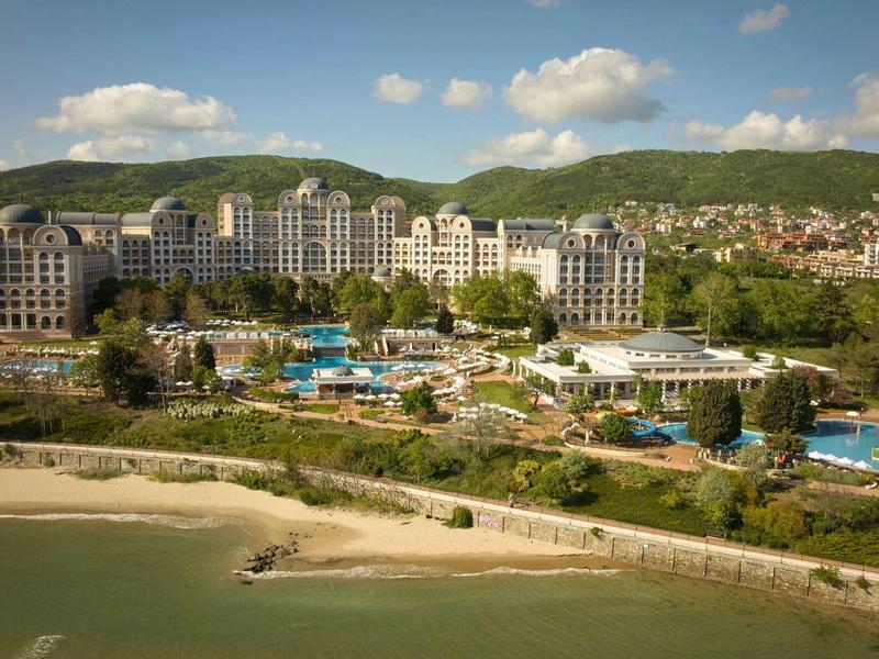 Large hotel resort with pools on the beach, surrounded by green hills and city.