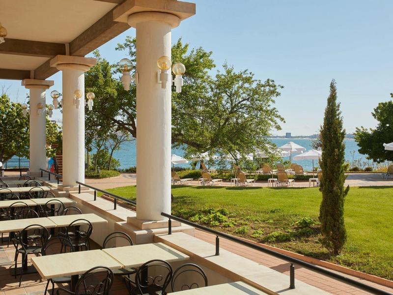 Covered terrace with tables and chairs overlooking garden, pool, and sea in the background.