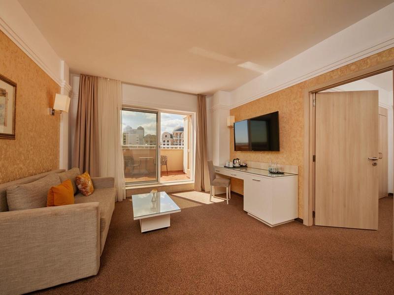 Bright hotel lounge with sofas, glass table, flat-screen TV, and balcony access.