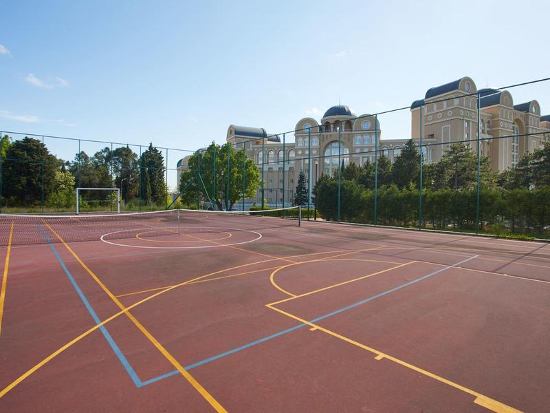 An empty tennis court with marked lines and trees in front of a large hotel building.