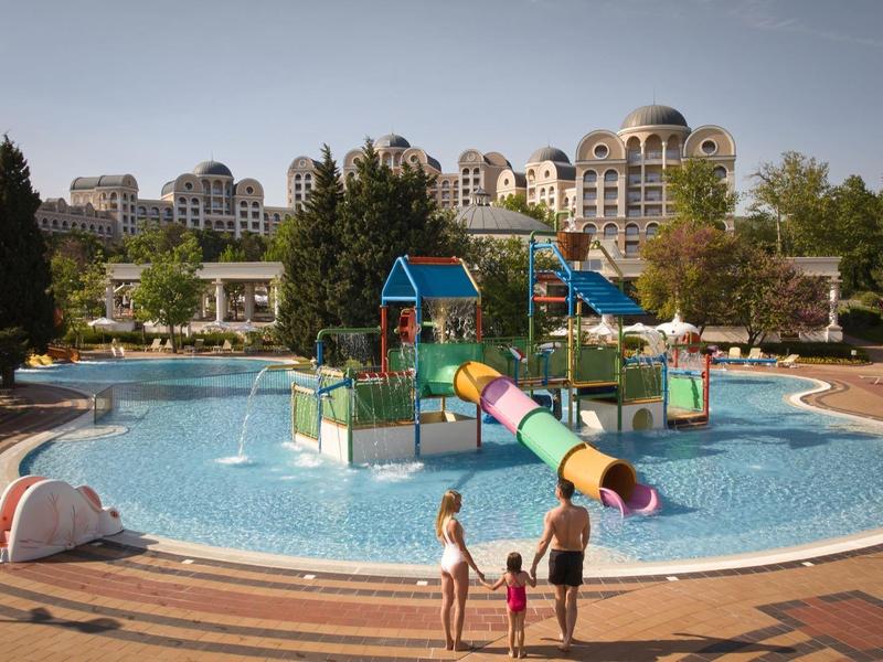 Family at a large outdoor pool with water slides and play structures in a resort.