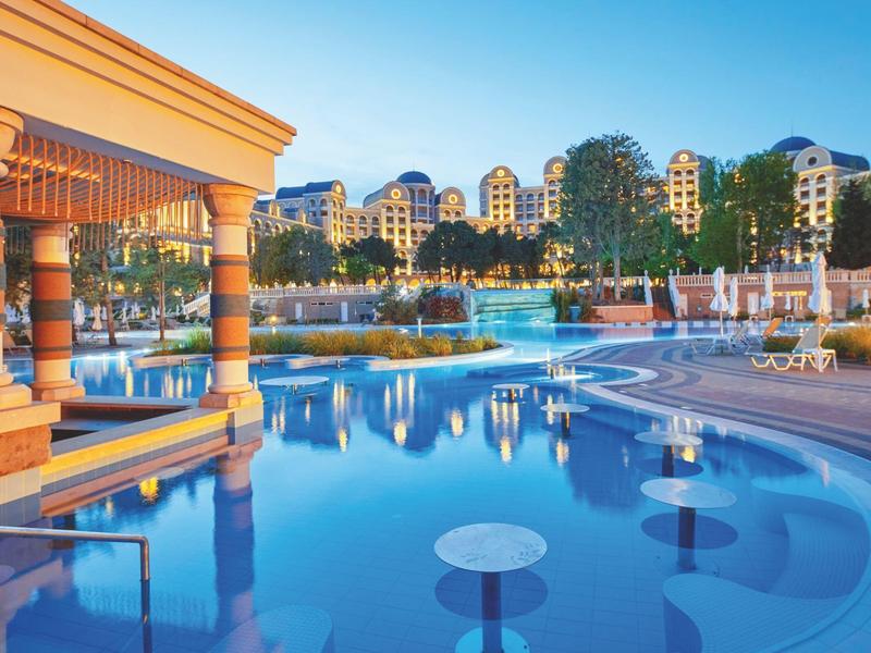 Luxury hotel pool with bar stools in water and Mediterranean-style buildings in background.