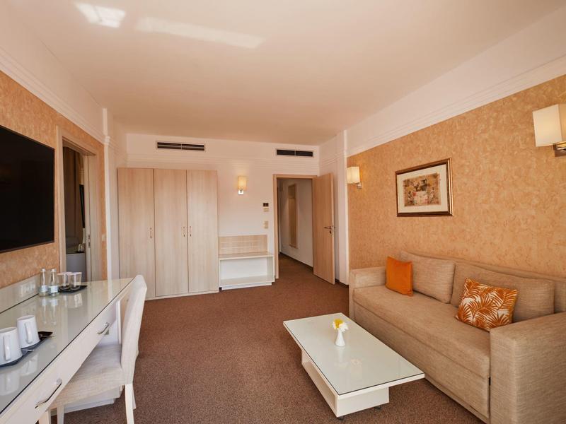 Modern hotel room with sofa, desk, and TV in warm tones.