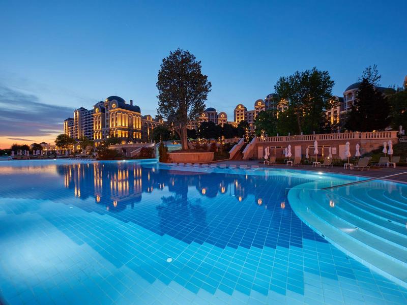 An illuminated outdoor pool in front of a hotel building at sunset.