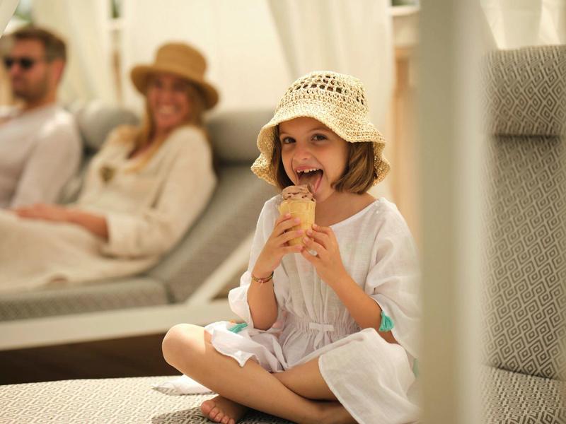 A smiling girl sits relaxed on a sun lounger holding an ice cream cone.