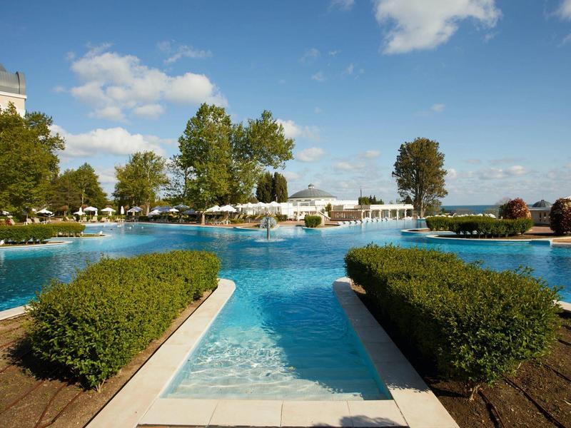 Wide pool with blue water basins and green hedges under a blue sky at a hotel resort.
