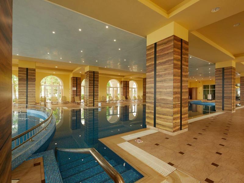 Indoor hotel area with swimming pool and mirrors, warm lighting, and light tiled floor.