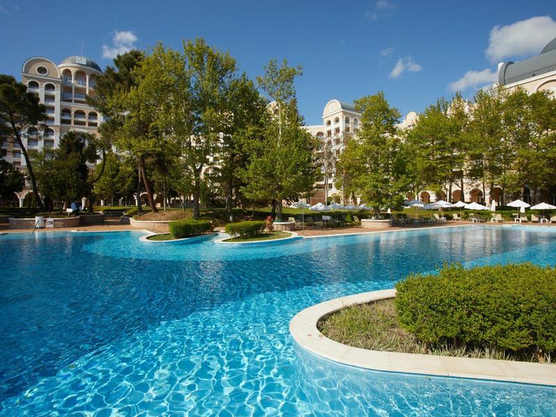 Large swimming pool with surrounding greenery and hotel buildings in the background