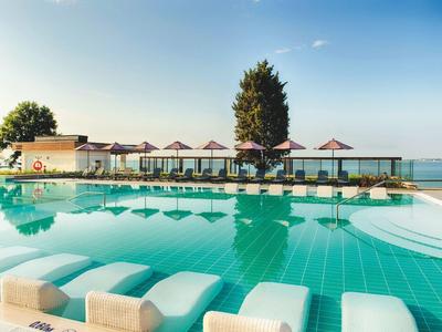 Großer Pool mit Liegen und Schirmen, dahinter Gebäude und Blick auf ruhiges Gewässer unter blauem Himmel.