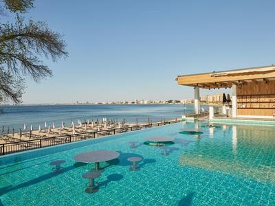 Infinity-Pool mit blauen Sitzgelegenheiten am Rand, Blick auf ruhiges Meer und bewachten Bootssteg im Sonnenschein.