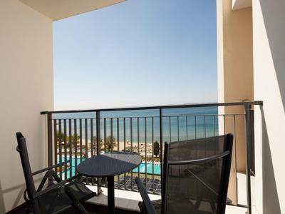 Balkon mit zwei Stühlen und kleinem Tisch, Blick auf Pool, Strand und ruhiges Meer bei klarem Himmel.