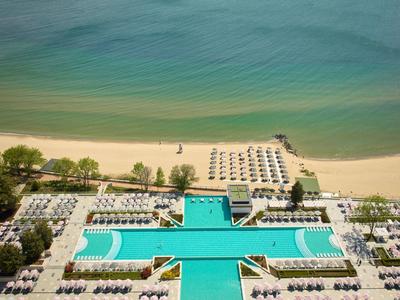 Großer Hotelpool mit Liegen an Sandstrand und grünem Meer im Hintergrund, von oben fotografiert.