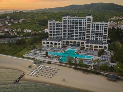 Großes Hotel mit Pool und Sonnenliegen am Strand vor Hügeln bei Sonnenuntergang.