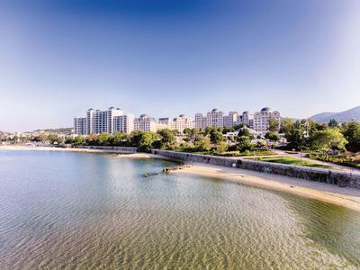 Küstenlinie mit Sandstrand, grünem Ufer und weißen Hochhäusern vor klarem blauem Himmel.
