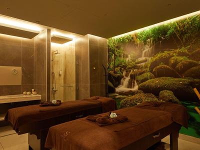 Gemütlicher Spa-Raum mit zwei Massageliegen, braunen Decken und Natur-Wasserfall-Wandbild.