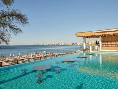 Pool mit blauen Tischen, Blick auf Meer und Strand mit Sonnenliegen unter blauem Himmel.