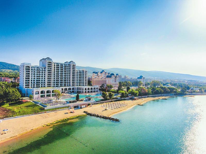 Große Hotelanlage am Strand mit Liegestühlen, klarem Wasser und Bergen im Hintergrund bei Sonnenschein.