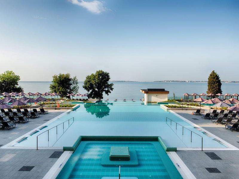 Moderner Infinity-Pool mit Liegestühlen, Blick auf ruhigen See und blauen Himmel.