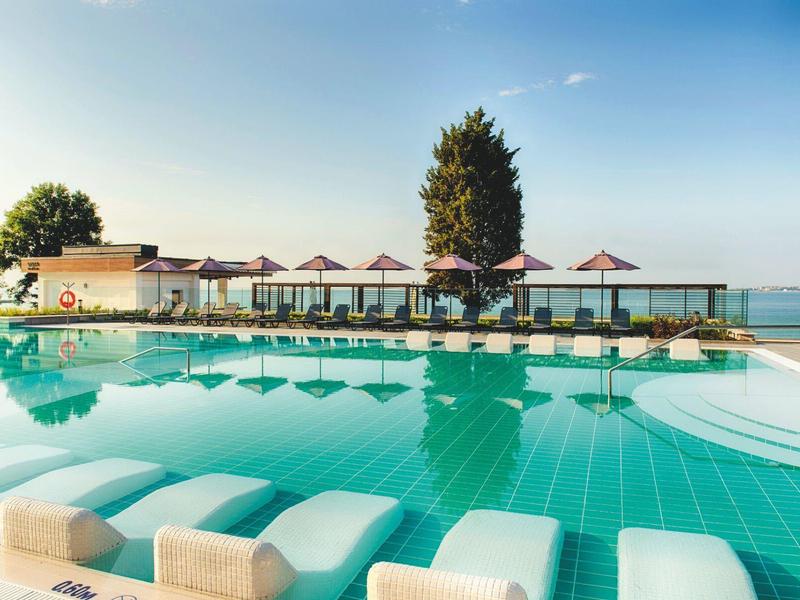 Großer Pool mit Liegen und Schirmen, dahinter Gebäude und Blick auf ruhiges Gewässer unter blauem Himmel.