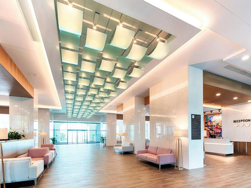 Großer heller Hotellobby mit moderner Deckenbeleuchtung, Sofas rechts und links, und Rezeption im Hintergrund.
