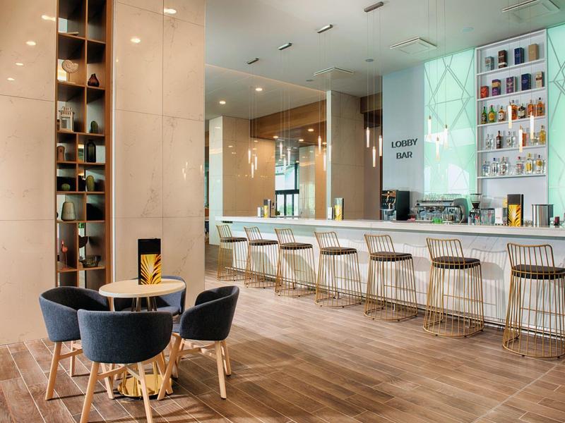 Moderner Barbereich mit heller Holztheke, weißen Sitzhockern und dunklen Sesseln vor Holzboden.