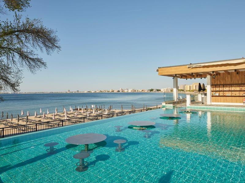 Infinity-Pool mit blauen Sitzgelegenheiten am Rand, Blick auf ruhiges Meer und bewachten Bootssteg im Sonnenschein.