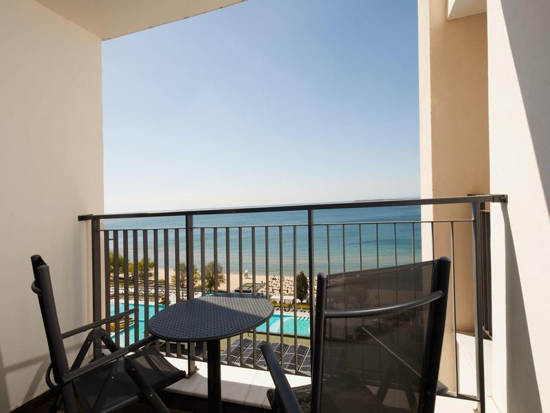 Balkon mit zwei Stühlen und kleinem Tisch, Blick auf Pool, Strand und ruhiges Meer bei klarem Himmel.