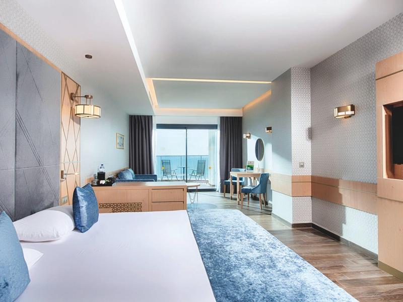 Modernes Hotelzimmer mit großem Bett, blauem Teppich, Schreibtisch und Balkon mit Aussicht.