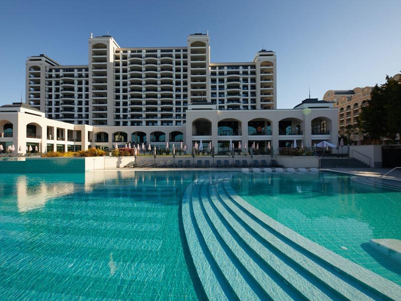 Großes Hotel mit weißen Arkaden und großem klaren Pool im Vordergrund unter blauem Himmel.