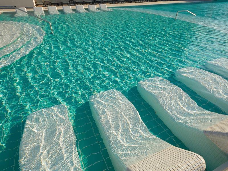 Schwimmbecken mit eingebauten weißen Liegen, blauem Wasser und Sonnenlichtreflexionen.