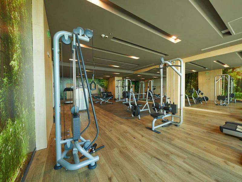 Moderner Fitnessraum mit Holzfußboden, verschiedenen Trainingsgeräten und großen Spiegeln.