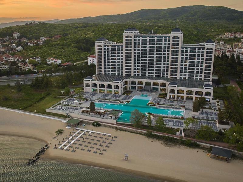 Großes Hotel mit Pool und Sonnenliegen am Strand vor Hügeln bei Sonnenuntergang.