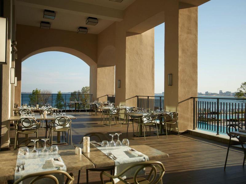 Terrassenrestaurant mit Holzboden, Tischen, Stühlen und Meerblick bei klarem Himmel.