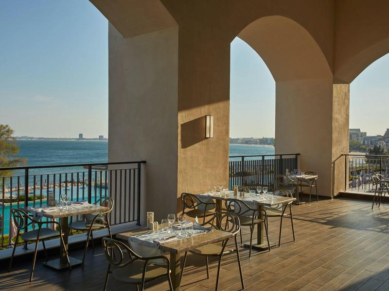 Balkon mit Stahlstühlen und Tischen, Blick aufs Meer und klare, sonnige Himmel.