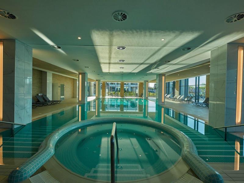 Moderner Innenpool mit hellem Holzdeck, großen Fenstern und integriertem Whirlpool im Vordergrund.