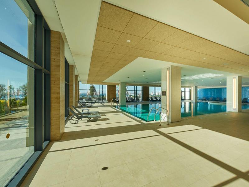 Moderner Innenpool mit großer Glasfront, Holzbalkendecke und Liegestühlen am Beckenrand.