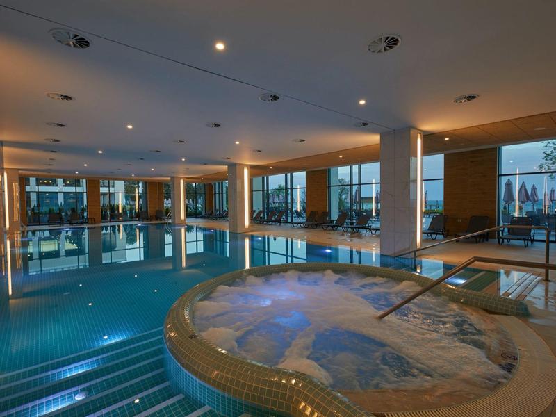 Innenraum mit großem Pool und rundem Whirlpool, moderne Beleuchtung und große Fenster.