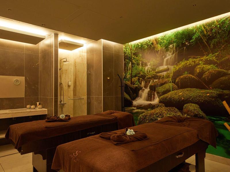 Gemütlicher Spa-Raum mit zwei Massageliegen, braunen Decken und Natur-Wasserfall-Wandbild.
