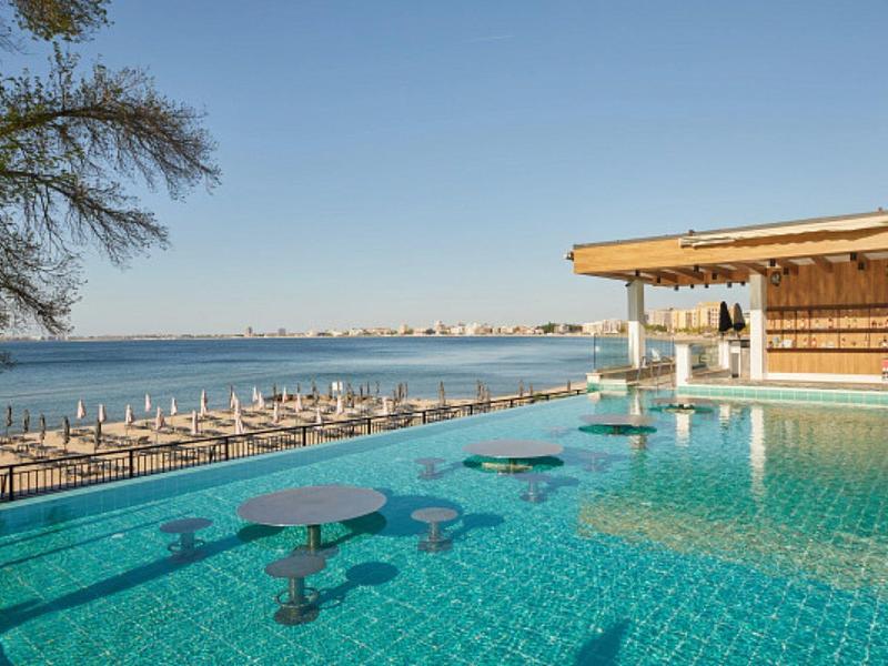 Pool mit blauen Tischen, Blick auf Meer und Strand mit Sonnenliegen unter blauem Himmel.