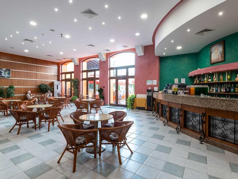 Bright hotel lounge area with wooden tables, chairs, and a bar on the side.